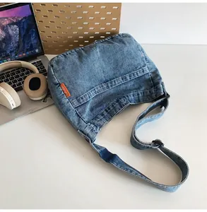 Blue denim crossbody bag with orange woven label on a workspace desk with laptop