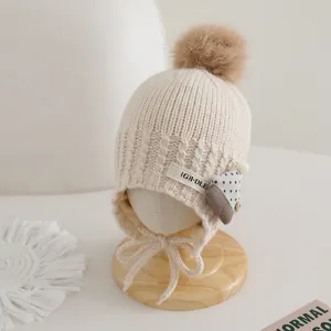 Detail of baby knit hat with brown pompom, earflaps, and strap on a wooden head stand