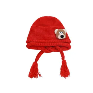 Red knit baby winter hat with cute bear patch, earflaps and festive tassels on white background