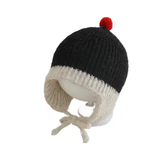 Gebreide kindermuts met oorkleppen in zwart-wit met rode pompon en strikbandjes