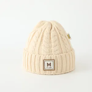Gros plan d'un bonnet en maille crème avec écusson lettre M et texture torsadée épaisse