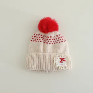 Detail of knit baby hat featuring red pom-pom, heart patterns and handmade lace patch