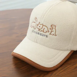 Detailaufnahme der beigen Fleece-Kinderkappe mit niedlicher Cartoon-Dinosaurier-Stickerei