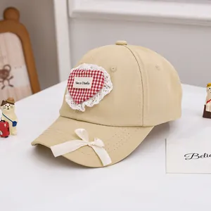 Détail d'une casquette enfant beige avec cœur à carreaux rouges et nœud en dentelle blanche