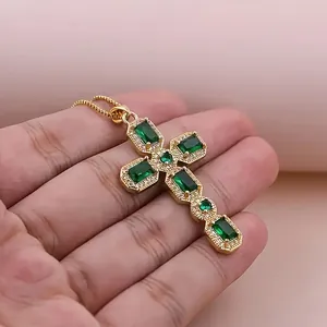 Hand holding a gold cross pendant necklace with green emerald stones and micro zircons