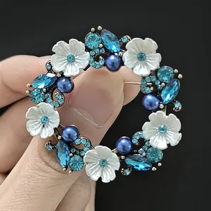Détail de la broche couronne de fleurs avec perles bleues et cristaux tenue à la main sur noir
