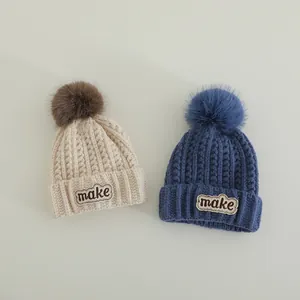 Close-up of knit winter hats with pom-poms and MAKE embroidery in cream and navy blue