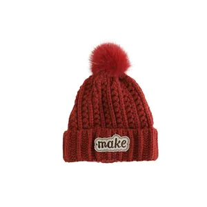 Winter children's knit hat in dark red with a large pompom and 'make' letter patch