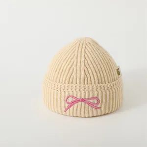Detailed view of cream ribbed knit beanie with pink bow embroidery and side logo tag
