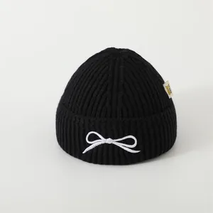 Close-up of black ribbed knit beanie featuring white bow embroidery and side fabric label