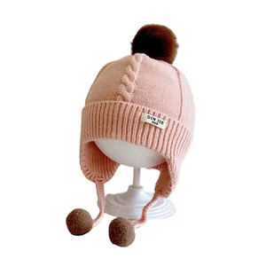 Pink knit baby earflap hat with brown pom poms and soft textured cable pattern