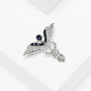 Détail d'une broche caducée argentée avec strass bleu foncé sur support géométrique blanc