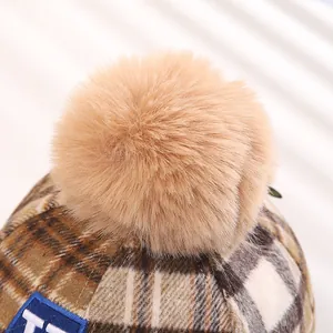 Close-up of winter checkered baby cap featuring a large fluffy faux fur pompom and letter patch