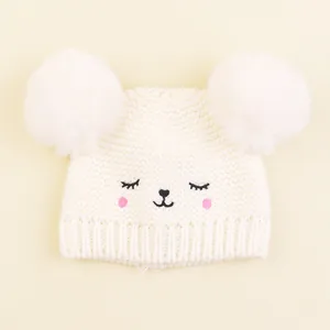 Close-up of white knit baby hat with two large faux fur pom poms and cute animal face embroidery