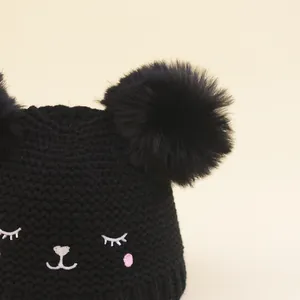 Kids black knit beanie featuring two large pom poms and cute cat face embroidery