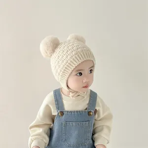 Detalle de gorro de punto para bebé con orejeras y pompones dobles en color beige suave