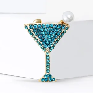 Broche verre à cocktail dorée avec strass bleus et perle blanche sur fond blanc