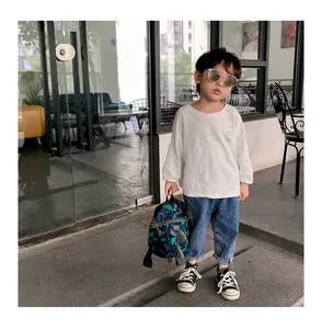 Outdoor shot of a toddler wearing a white long-sleeve tee with denim jeans and dinosaur bag