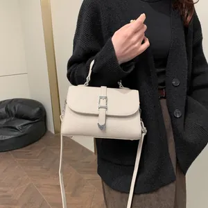 Sac bandoulière crème en cuir grainé avec boucle cœur porté sur un gilet noir