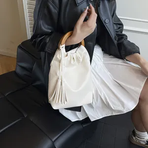 Elegante bolso de cubo blanco crema con asa de madera y borlas sobre banco de cuero
