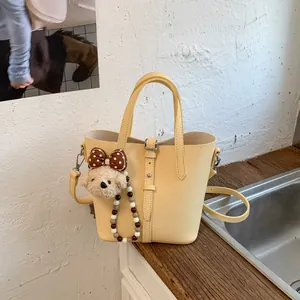 Pale yellow leather bucket bag with top handle and cute plush bear charm on white wall