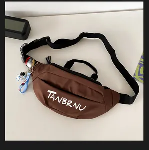Brown nylon chest bag with TANBRNU text, black strap, and astronaut charm on a desk