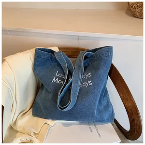 Detailed close-up of white 'Less More Mondays' embroidery on blue denim tote bag surface