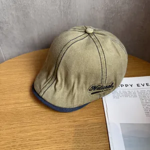 Detail of khaki cotton newsboy cap featuring navy contrast stitching and Natural embroidery