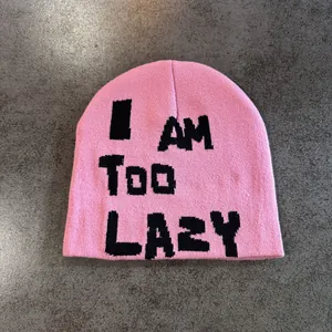 Detail of pink knitted beanie with black I AM TOO LAZY text on textured grey background