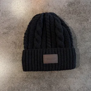 Detail van zwarte kabelgebreide muts met warme oorkleppen en een bruine lederen patch