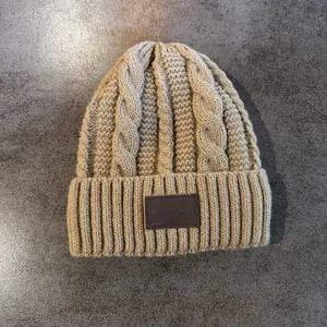 Detail van een beige kabelgebreide muts met een bruine leren patch-accent