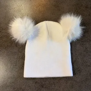 Detalle de gorro de punto blanco con dos pompones de piel sintética sobre mármol oscuro