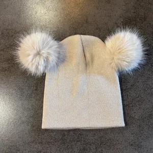 Detalle del gorro de punto beige con dos pompones de piel sintética sobre fondo piedra