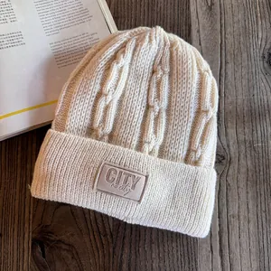 Detail of cream knit beanie featuring thick chain texture and leather brand label on a desk