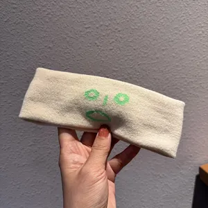 Détail du bandeau large en tricot crème avec un motif de visage brodé en vert