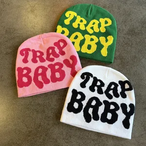 Three TRAP BABY jacquard knit beanies in pink, green, and white on a textured background