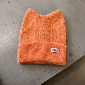 Detailansicht der orangefarbenen Katzenohr-Mütze mit flauschiger Textur und Webetikett