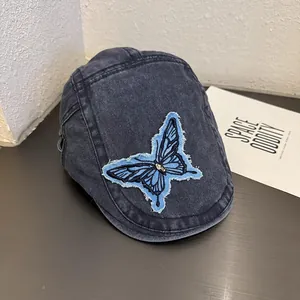 Close-up of blue butterfly embroidery on washed denim newsboy cap with distressed edges