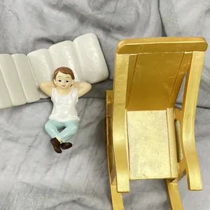 Miniature resin boy figure lying down next to a vintage gold rocking chair for dollhouse decor