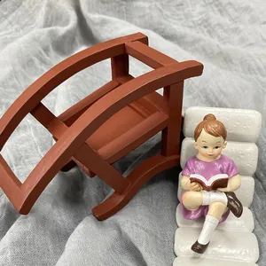 Miniature figurine of a girl reading on a white chair next to a tiny dark wood arch bridge