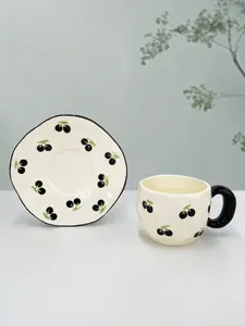 Detail of handmade ceramic coffee cup and saucer with black cherry patterns on milky white