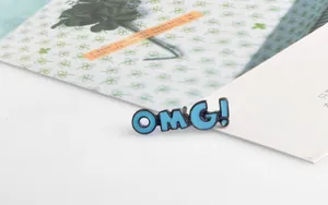 Detailed view of an 'OMG!' blue enamel brooch on a stylish decorative card and envelope
