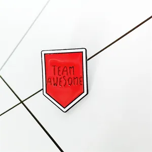 Dettaglio di una spilla smaltata rossa a forma di scudo con testo TEAM AWESOME