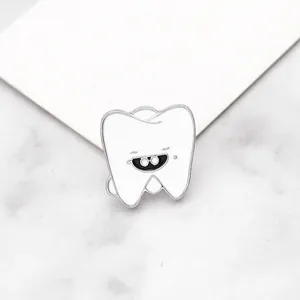Détail d'un pin's en émail blanc en forme de dent avec un visage souriant et des bords argentés