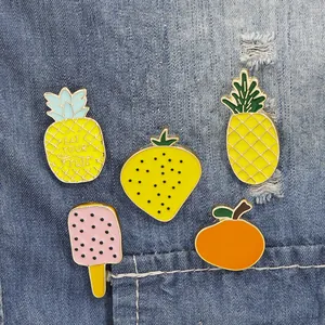 Five assorted colorful cartoon enamel fruit pins on denim fabric including pineapple and orange