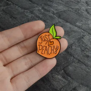 Gros plan détaillé d'un pin's en émail pêche avec texte JUST PEACHY tenu dans la main