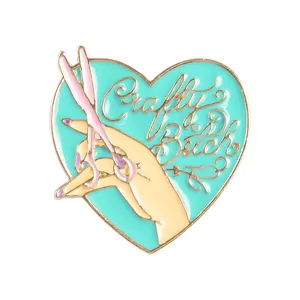 Herzförmige Emaille-Anstecknadel mit Crafty Bitch Text und rosa Schere auf blauem Grund
