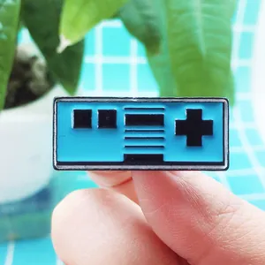Close-up of a blue retro game controller enamel pin held by fingertips showing the small size