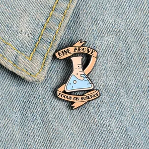 Detail of beaker enamel pin with Rise Above and Focus on Science text on denim jacket