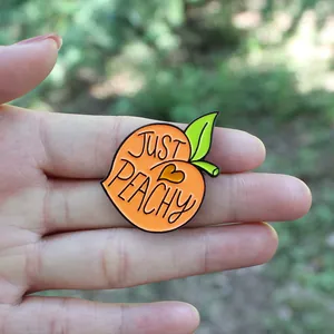Détail du pin's pêche en émail avec slogan 'JUST PEACHY' tenu dans la main pour taille